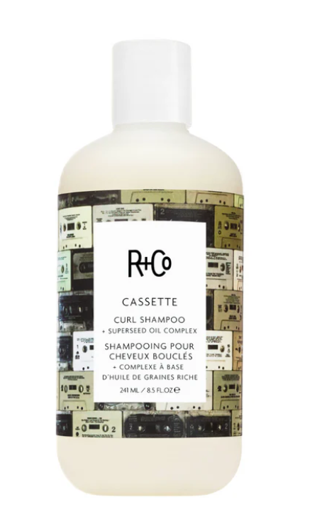 A 240 mL bottle of R+Co Cassette Curl Shampoo, featuring a white cap and a label with a retro cassette tape collage pattern on a clean white background.